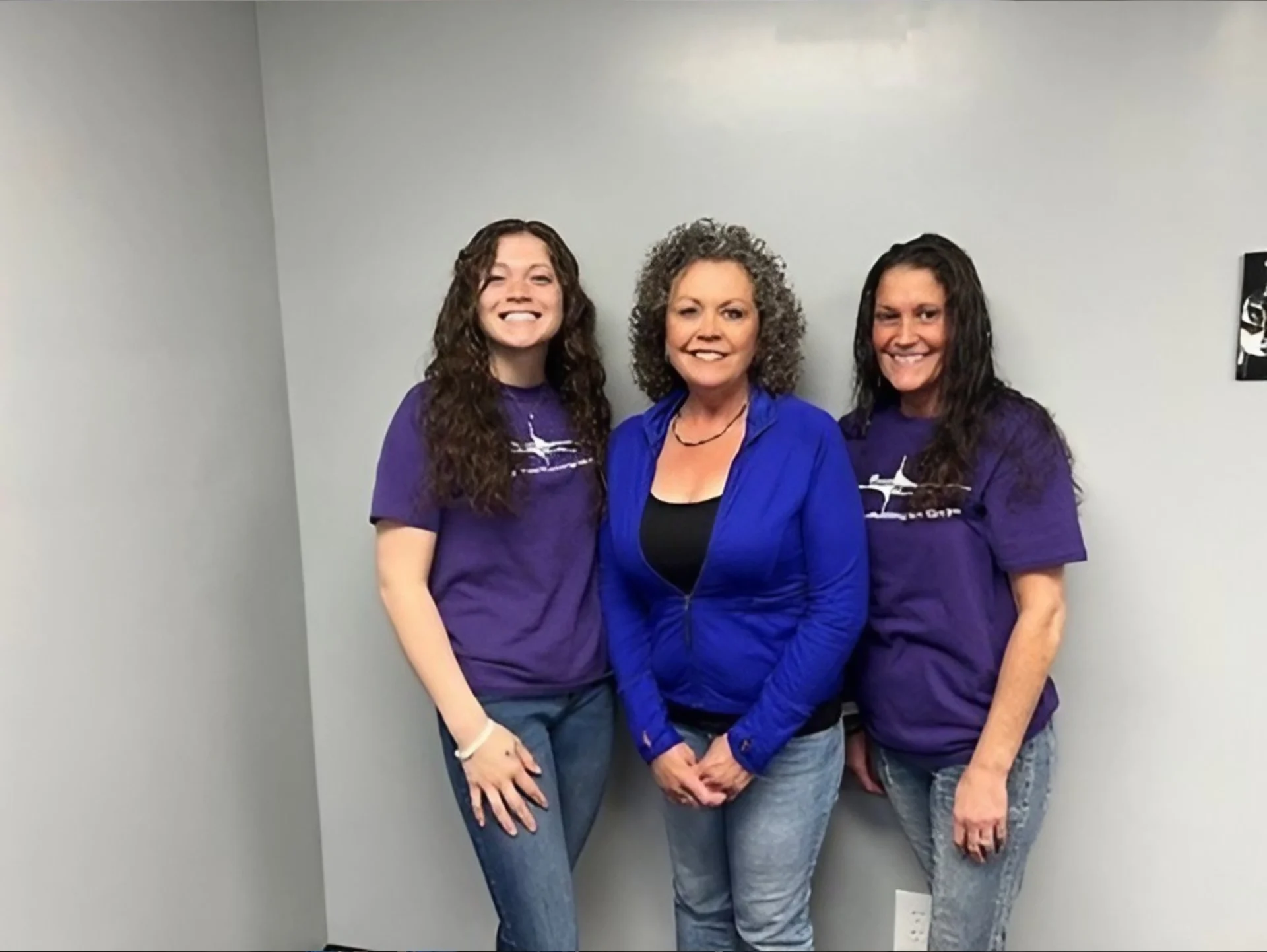 Three women smiling, two in purple shirts and one in a blue sweater.