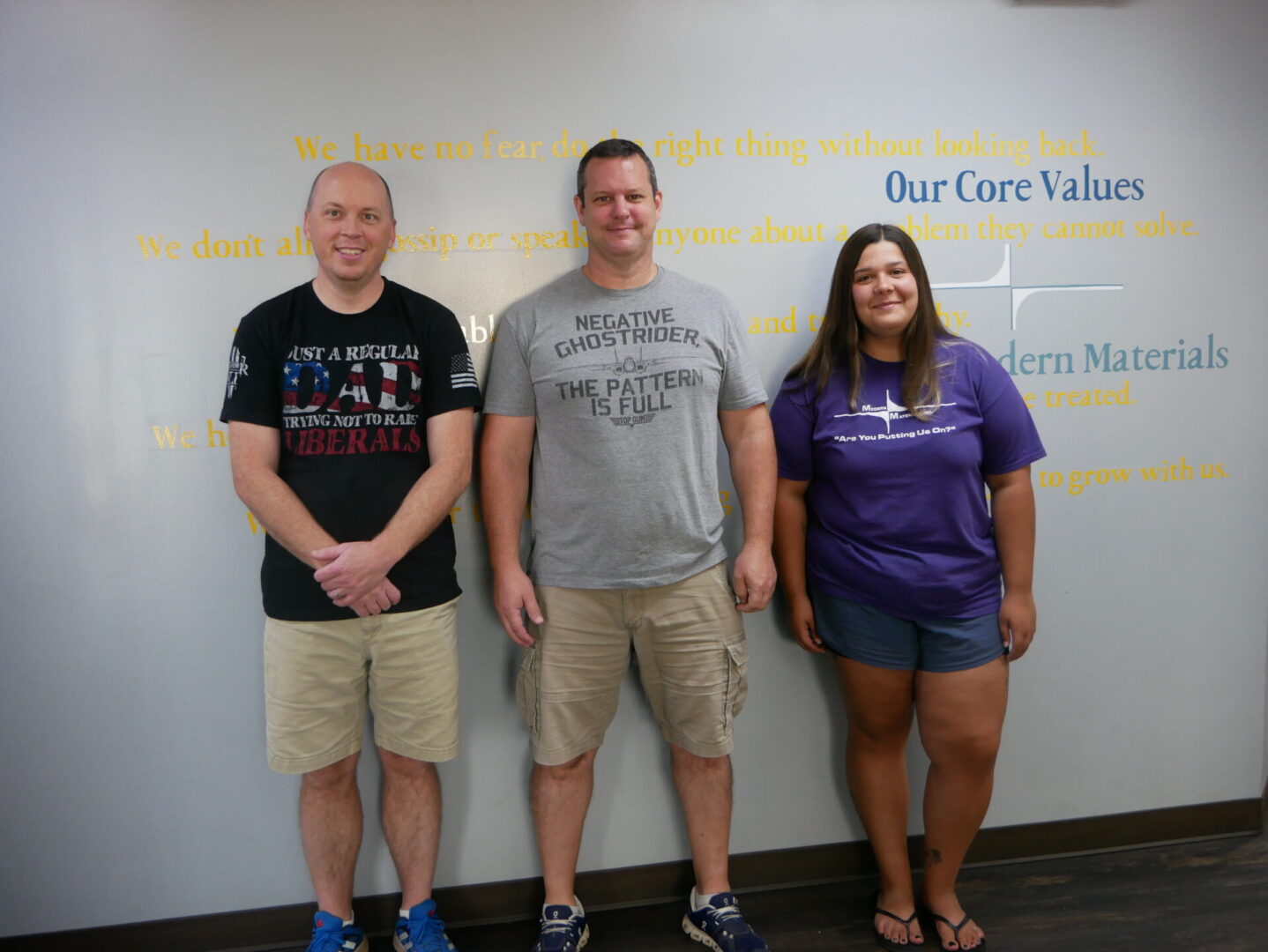 Three people smiling and standing indoors in front of a wall with text.